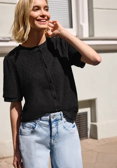 Vrouw draagt zwart, knoopsluitend T-shirt met korte mouwen en blauwe jeans voor een gevel.